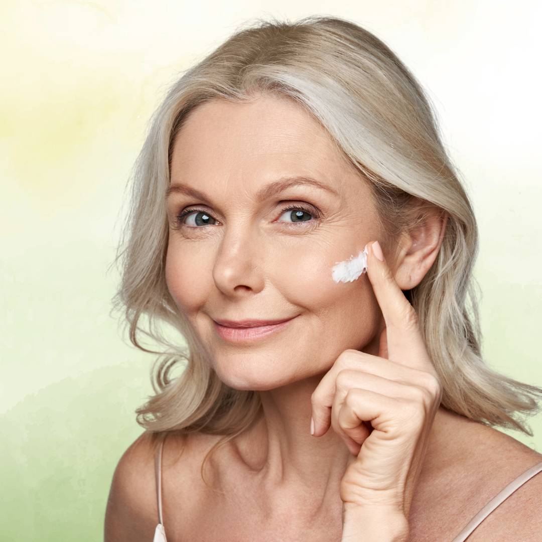 Mulher de meia idade e serena, com fundo verde claro, aplicando suavemente creme anti-idade no rosto, evidenciando um momento de cuidado e bem-estar natural.