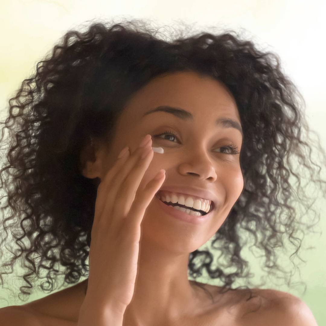 Mulher jovem e radiante, com fundo verde claro, aplicando suavemente creme no contorno dos olhos, evidenciando um momento de cuidado e bem-estar natural.