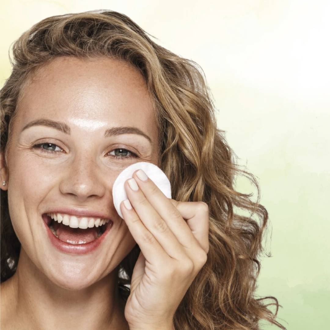 Mulher jovem e radiante, com fundo verde claro, aplicando suavemente o tónico com um disco de algodão no rosto, evidenciando um momento de cuidado e bem-estar natural.